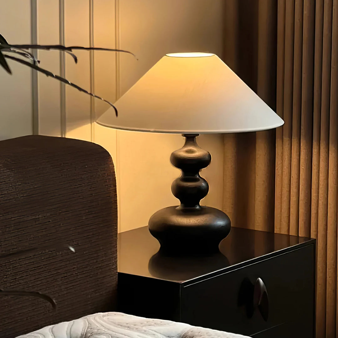 A beautifully lit bedroom scene with a Warm Ambient Gourd Lamp placed on a glossy black bedside table. The lamp features a dark wooden base with a sculptural form and a classic white shade that diffuses soft light, enhancing the cozy atmosphere. The warm glow complements the natural wood textures and vertical blinds in the background, adding a touch of elegance and tranquility to the space.