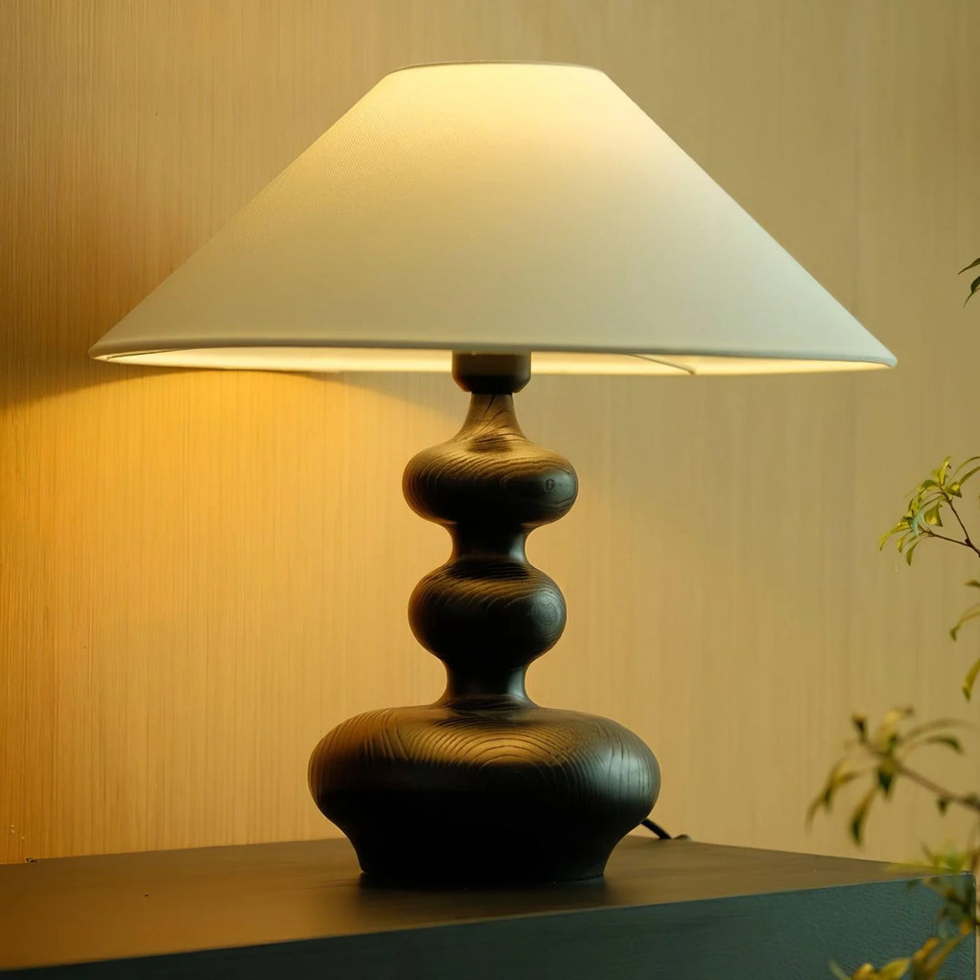 A close-up view of a Warm Wooden Gourd Table Lamp featuring a unique, sculptural base with flowing curves in a dark wood finish, topped with a large, bright white shade. The lamp casts a gentle glow, enhancing the warm ambiance against a soft-lit wooden backdrop, ideal for creating a cozy and inviting space.