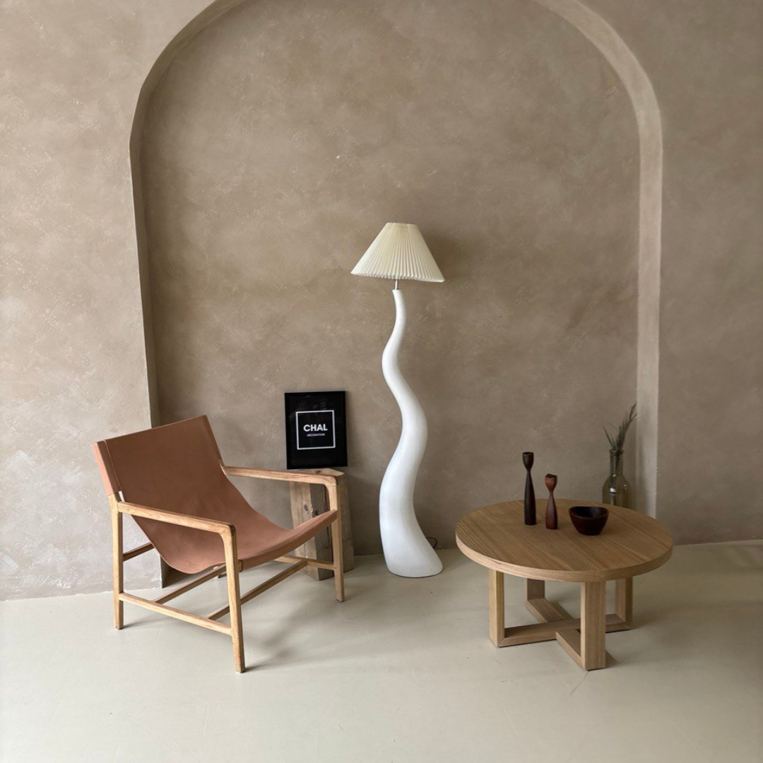 A wavy white floor lamp with a pleated shade, styled in a minimalist living room with an arched alcove, a leather chair, and a wooden coffee table featuring decorative accents.