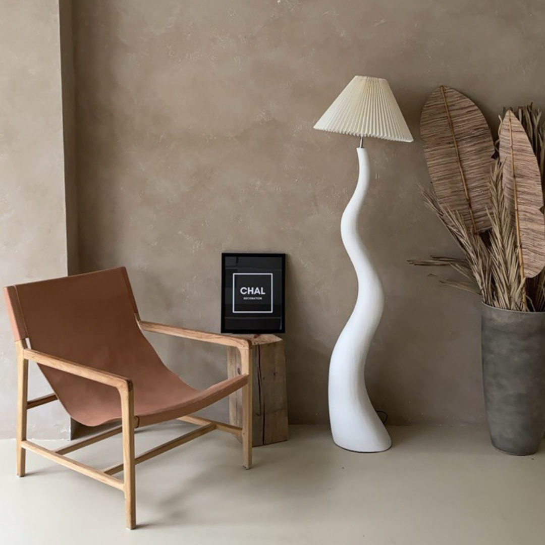 A wavy white floor lamp with a pleated shade, placed in a boho-inspired living room featuring a leather accent chair, neutral decor, and dried palm leaves in a large vase.