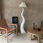 Load image into Gallery viewer, The wavy floor lamp in a stylish living space with a natural wood coffee table, leather chair, and minimalist decor elements on a neutral-toned background.
