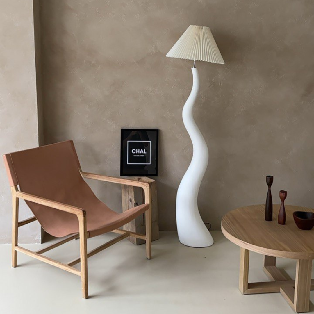 A sculptural floor lamp with a wavy white base and a pleated shade, styled in a modern living room with neutral tones, a leather chair, and a wooden coffee table.