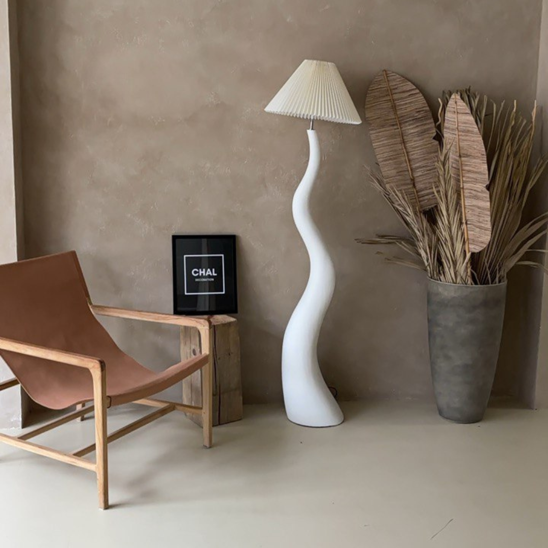 The wavy floor lamp placed in a cozy, minimalist living space with a neutral-toned wall, a wooden chair, and decorative dried plants in a vase.