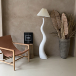 Load image into Gallery viewer, The wavy floor lamp placed in a cozy, minimalist living space with a neutral-toned wall, a wooden chair, and decorative dried plants in a vase.
