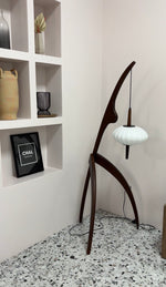 Load image into Gallery viewer, The wooden mantis floor lamp placed in a modern corner with decorative shelves, a framed art piece, and terrazzo flooring, highlighting its unique design and craftsmanship.
