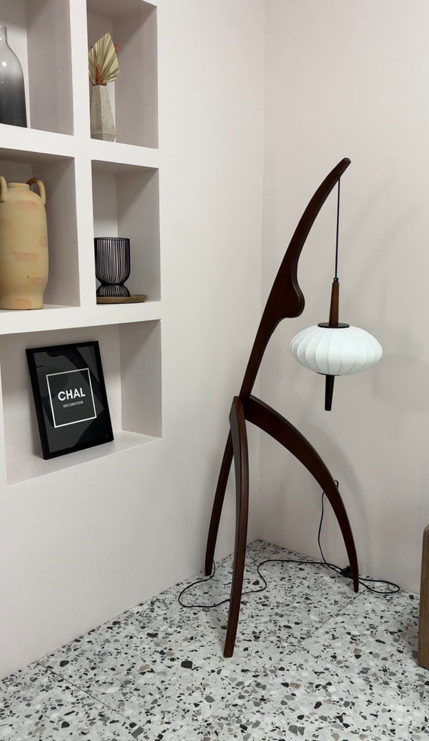 The wooden mantis floor lamp placed in a modern corner with decorative shelves, a framed art piece, and terrazzo flooring, highlighting its unique design and craftsmanship.