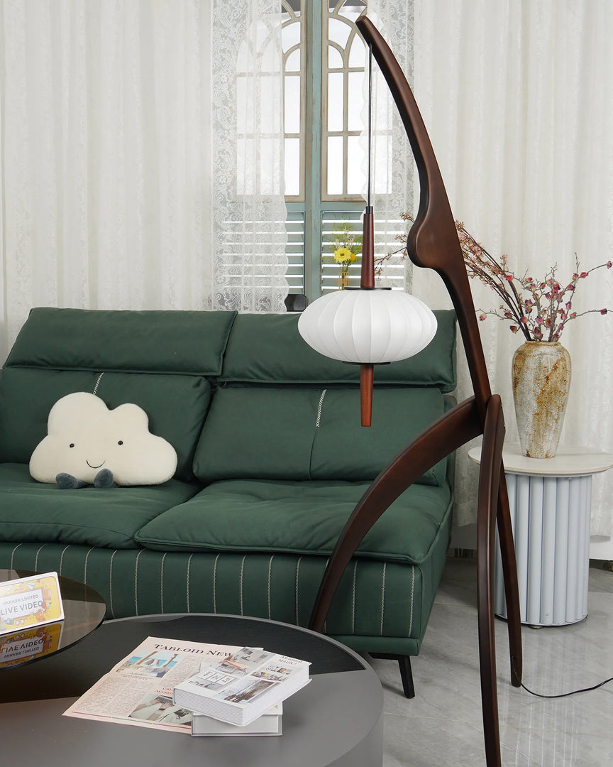 The wooden mantis floor lamp in a cozy living room with a green sofa, a cloud-shaped pillow, a round coffee table, and a vase with decorative flowers near a window.