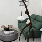 Load image into Gallery viewer, The wooden mantis floor lamp placed in a modern living room setting with a green sofa, a round coffee table, and contemporary decor elements.
