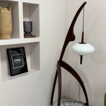 Load image into Gallery viewer, A unique wooden floor lamp with a mantis-inspired design, featuring a curved frame and a hanging white lantern-style lampshade, placed in a modern minimalist corner with decorative shelves.
