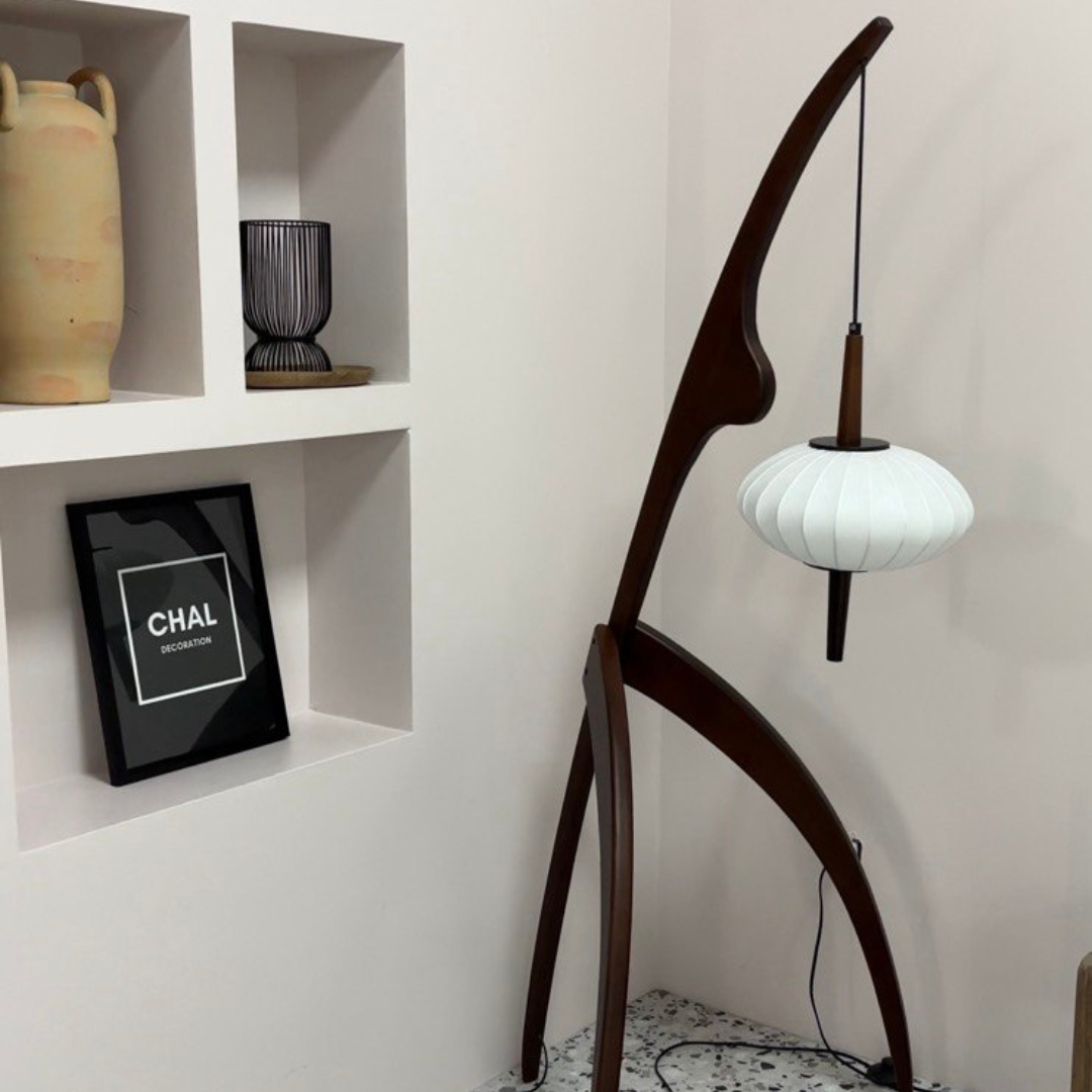 A unique wooden floor lamp with a mantis-inspired design, featuring a curved frame and a hanging white lantern-style lampshade, placed in a modern minimalist corner with decorative shelves.